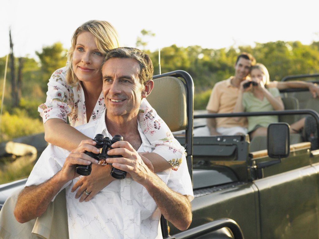 Two couples on safari.  One on the hood of the safari jeep, the second couple in the jeep with both couples holding binoculars and smiling